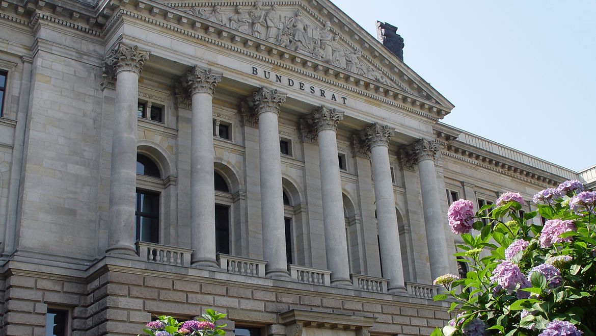 Das Gebäude des Bundesrats. Das Bild zeigt die Vorderseite eines großen alten Gebäudes. 6 Säulen zieren den Eingangsbereich, darüber steht geschrieben BUNDESRAT, Im Vordergrund sind Blumen abgebildet (Hortensien).