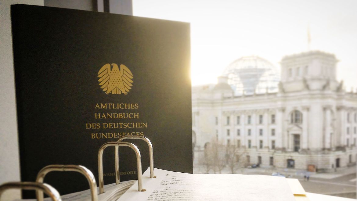 das Amtliche Handbuch des Deutschen Bundestages vor einem Fenster, im Hintergrund das Reichstagsgebäude mit der Kuppel Amtliches Handbuch des Deutschen Bundestages