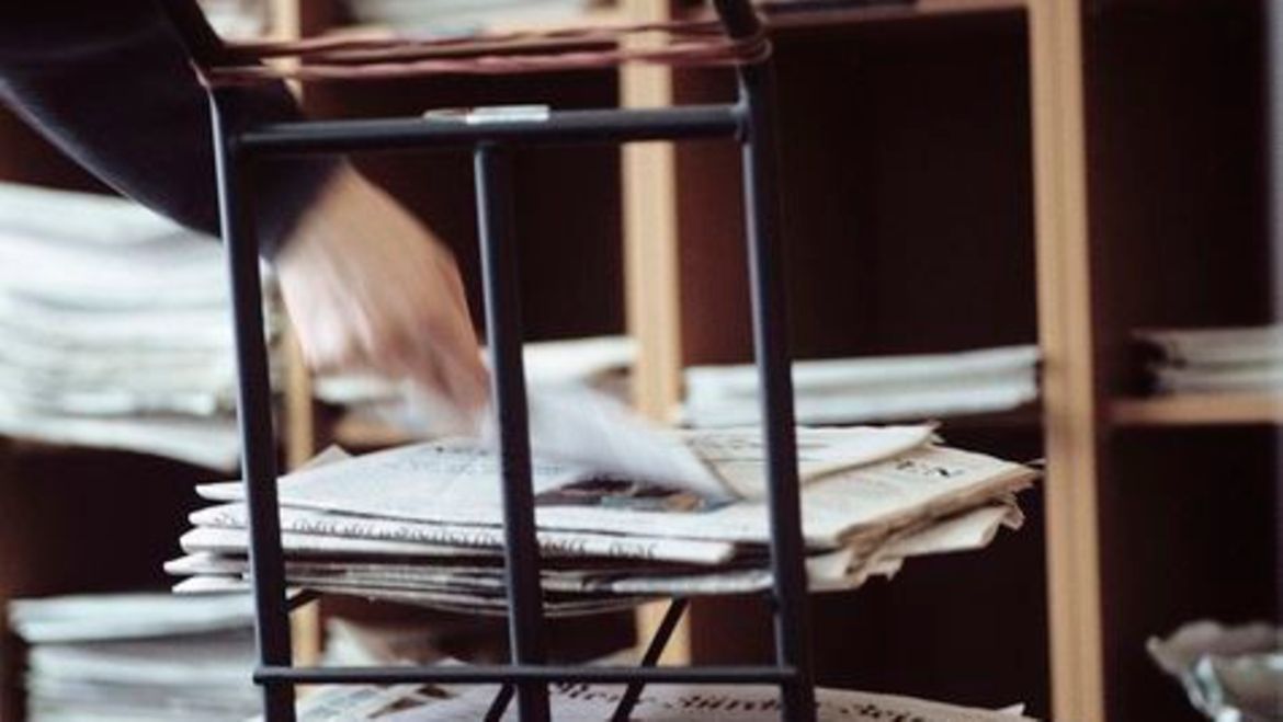 Die Pressedokumentation wertet Zeitungen aus. Symbolbild mit einer Hand, die eine Zeitung auf einen Stapel Zeitungen legt.