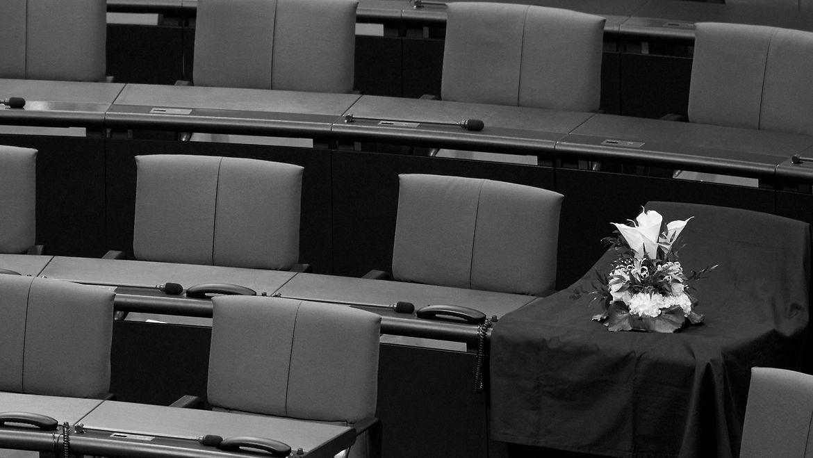 Ein digitales Gedenkbuch erinnert an die verstorbenen Abgeordneten des Bundestages. Schwarz-weiß-Aufnahme der leeren Sitzreichen im Plenarsaal des Bundestages mit einem Trauergesteck auf einem Platz